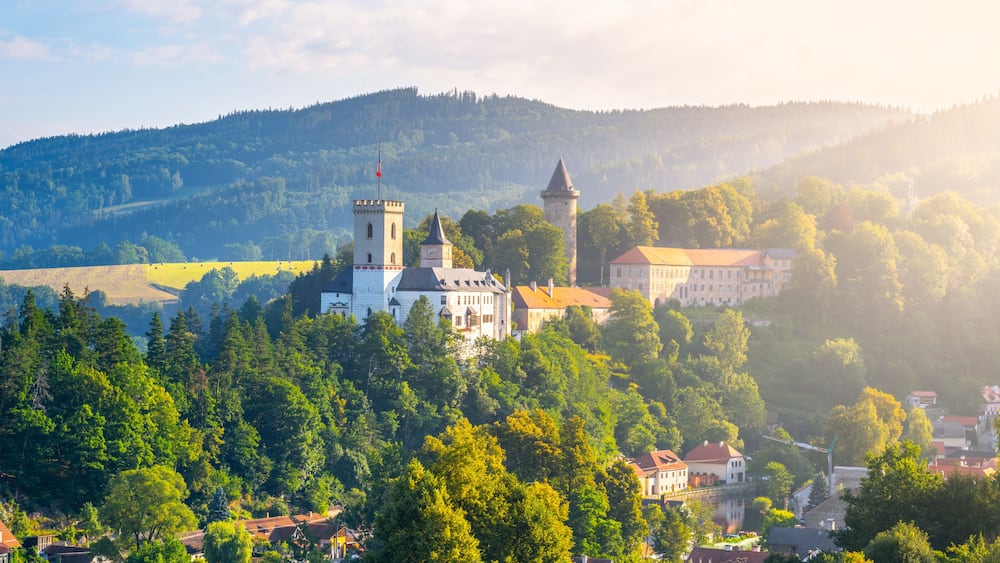 A castle surrounded by forested hills, a church with a clock tower, and a river.
