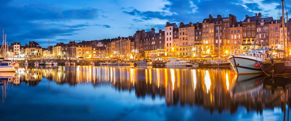 A waterfront cityscape at dusk with illuminated buildings reflecting on the water.