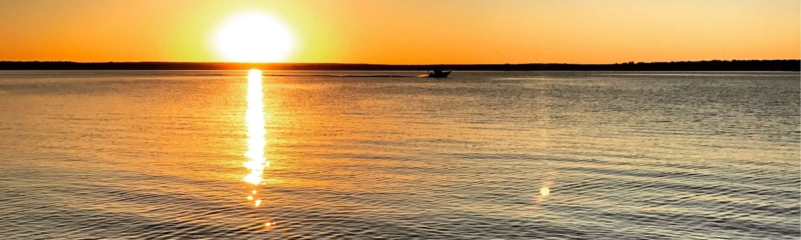Un atardecer sobre una masa de agua tranquila, con un barco a lo lejos y un litoral rocoso en primer plano.