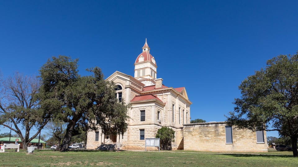 A historic building with a red dome and a clock tower, surrounded by trees and a grassy field.
