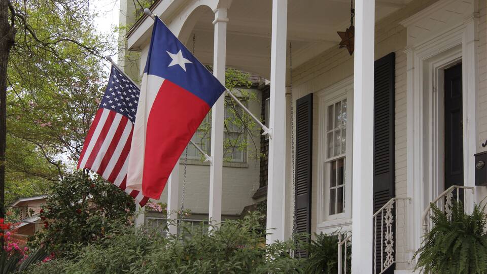A porch with two flags, one American and one Texas state flag, and a white building with black shutters.