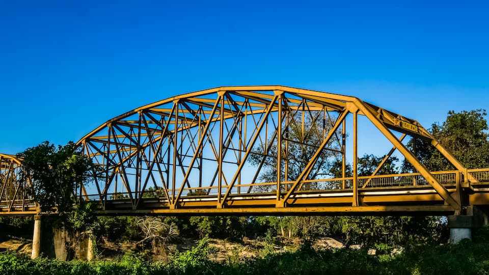 A large steel bridge with a curved arch design, spanning over a river with dense greenery and trees in the background.