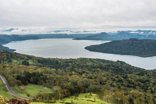 Ein See, umgeben von bewaldeten Hügeln, eine kurvenreiche Straße und entfernte Berge.