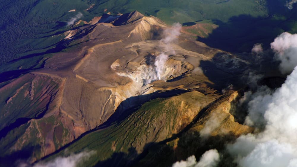 A volcanic landscape with a central crater emitting smoke, surrounded by green forested areas and rugged terrain.
