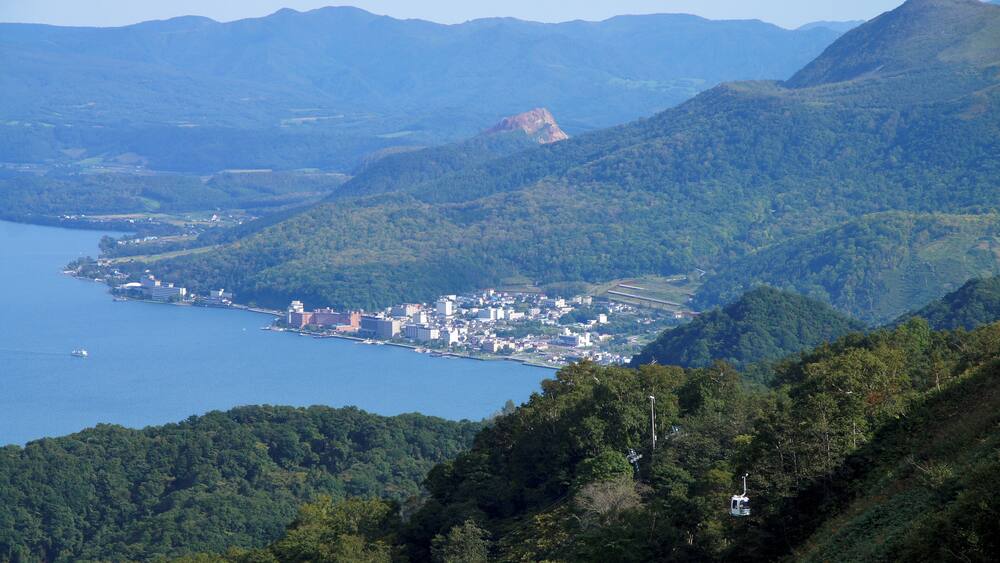 A coastal town nestled between mountains, with a large body of water to the left and dense forested hills.