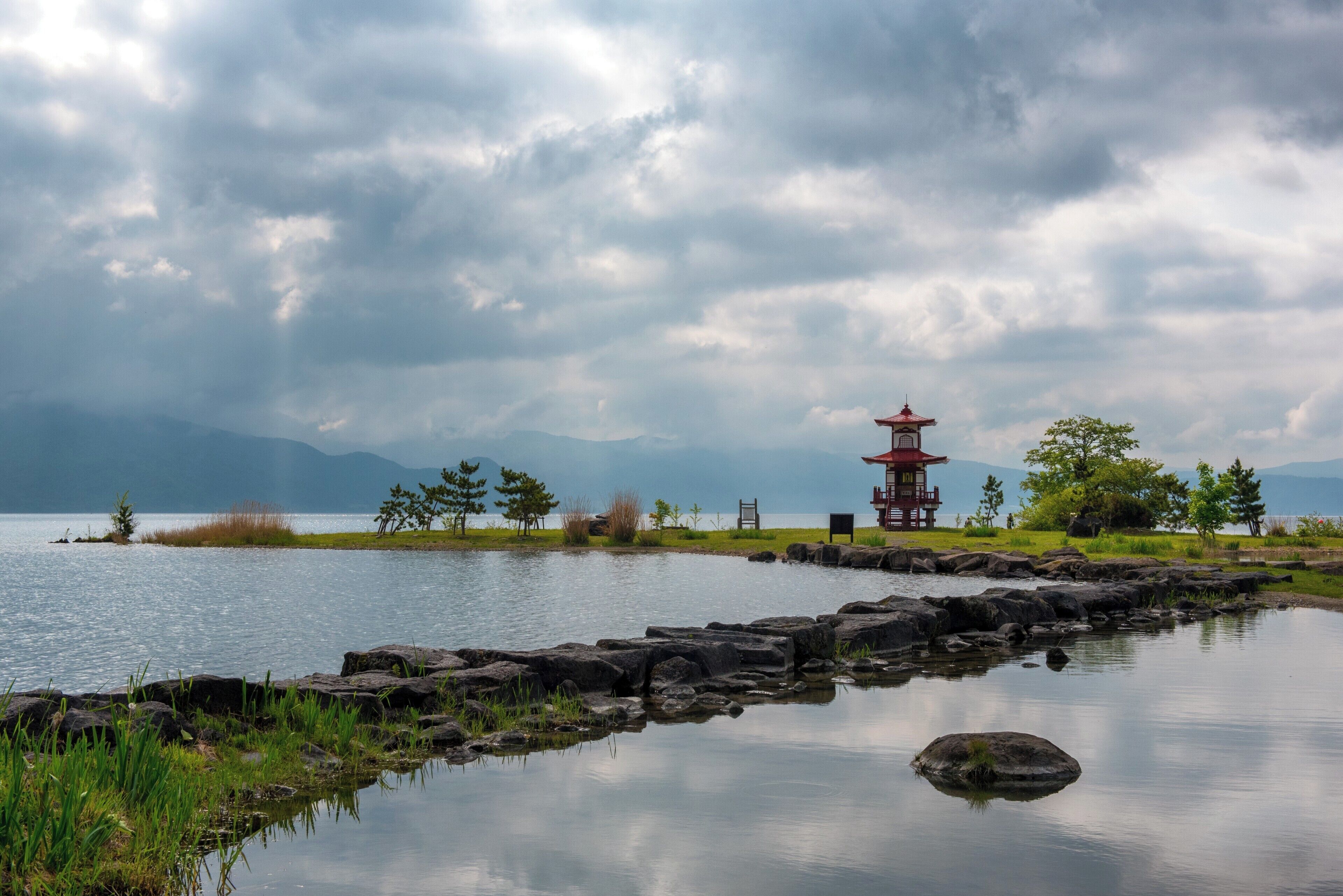 A tranquil lakeside scene with a traditional red pagoda, rocky shoreline, and distant mountains under a cloudy sky.