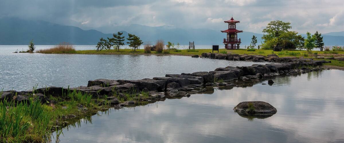 A tranquil lakeside scene with a traditional red pagoda, rocky shoreline, and distant mountains under a cloudy sky.