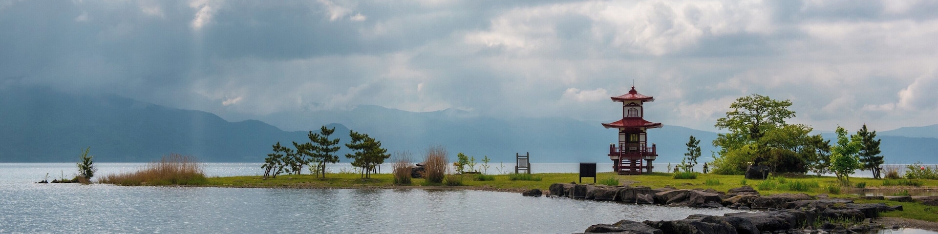 A tranquil lakeside scene with a traditional red pagoda, rocky shoreline, and distant mountains under a cloudy sky.
