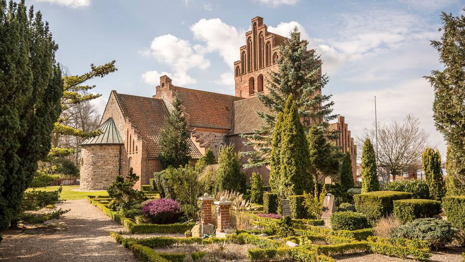 A historic brick building with a tower, surrounded by well-manicured gardens and tall trees.