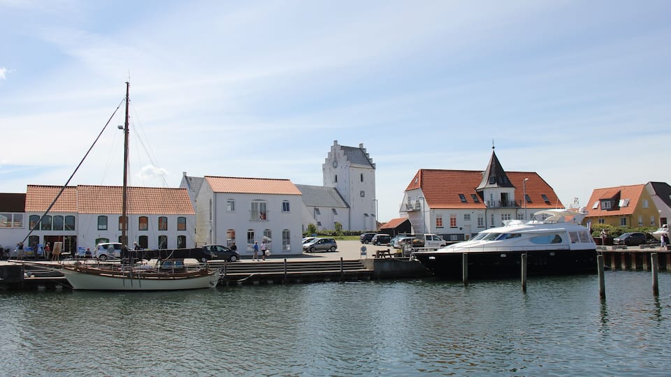 A harbor with a boat docked, buildings with red roofs, and a church tower in the background.