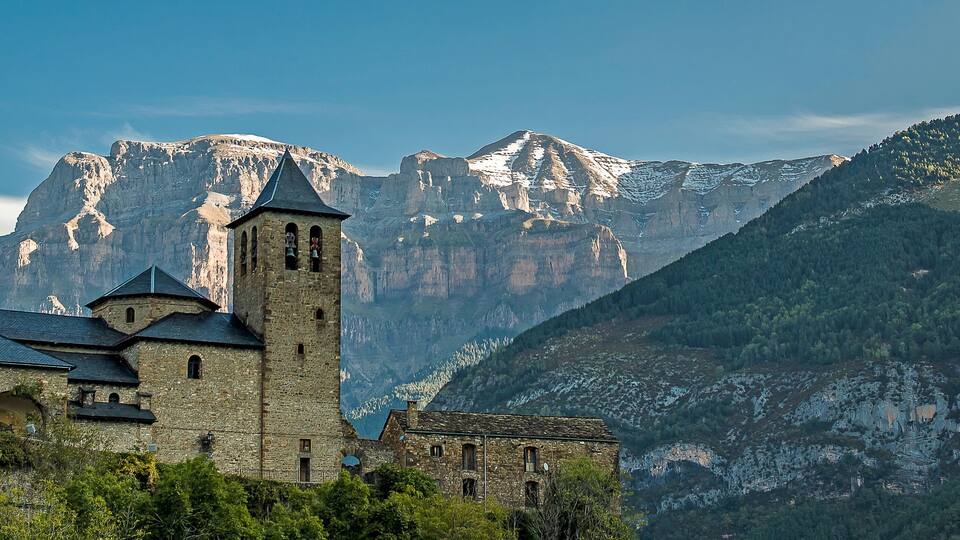 A historic church with a bell tower, set against a backdrop of snow-capped mountains and lush greenery.