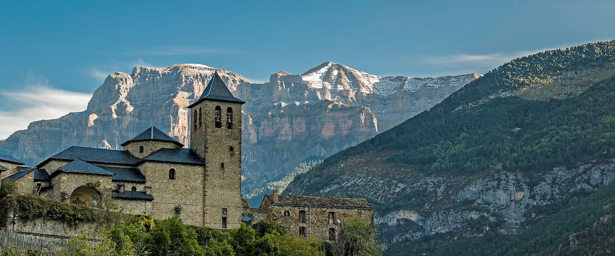 A historic church with a bell tower, set against a backdrop of snow-capped mountains and lush greenery.