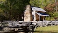 A rustic log cabin with a stone chimney, surrounded by a wooden fence and a stone wall.