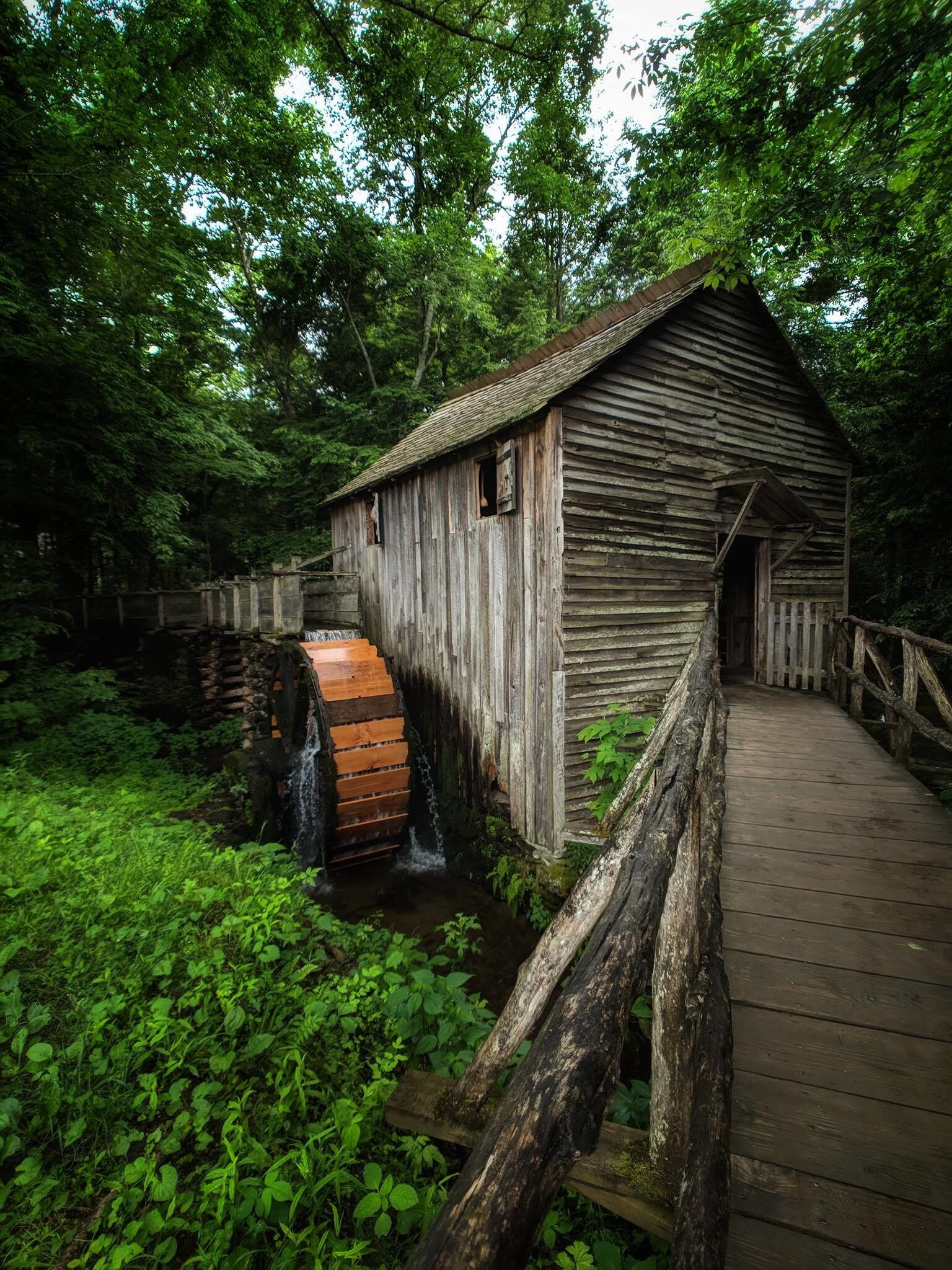 Een houten watermolen met een houten looppad ernaartoe, omgeven door weelderige begroeiing en een kleine waterval.