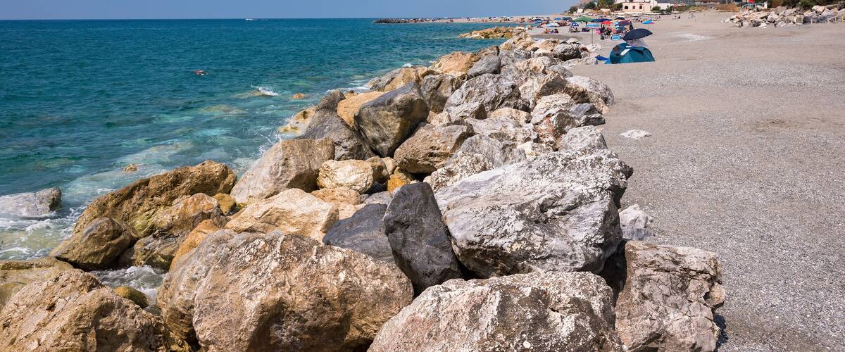 A rocky shoreline with clear blue water, a sandy beach, and a distant mountain range.