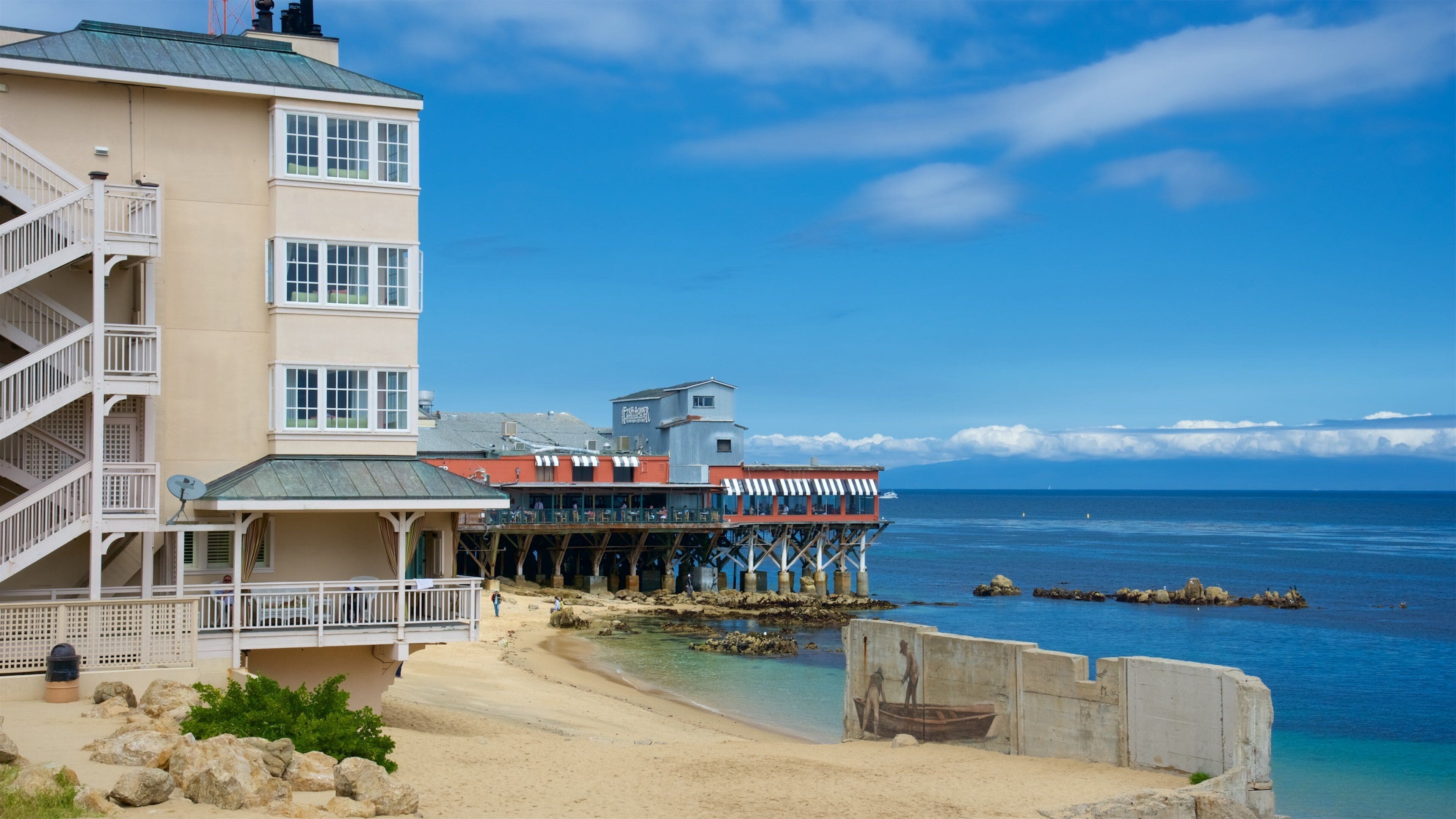 A coastal building with a balcony overlooking the sea, a pier extending into the water, and a sandy beach.
