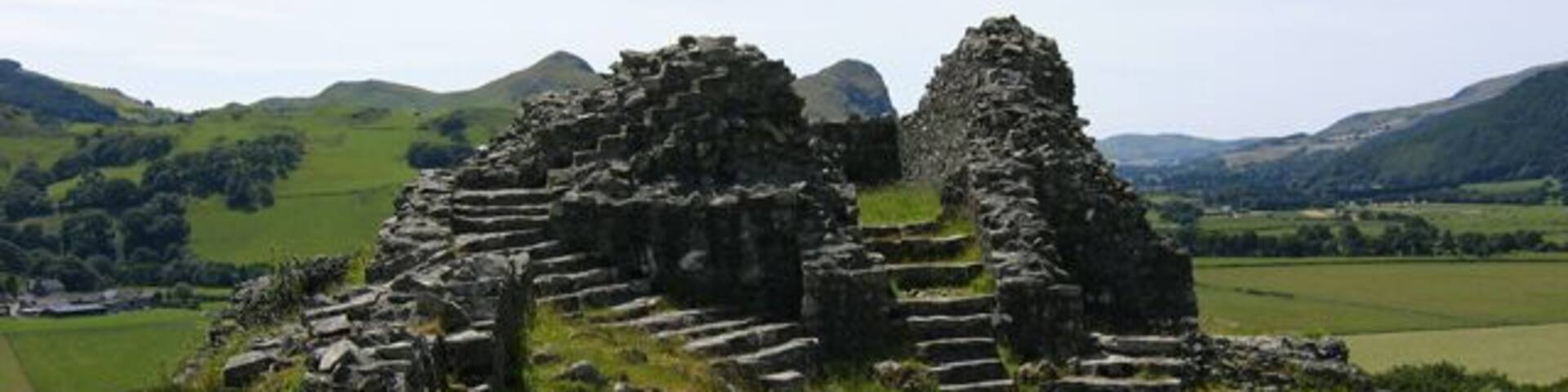 Ancient stone ruins with stepped structures and a large stone wall, set against a backdrop of rolling hills and a clear sky.