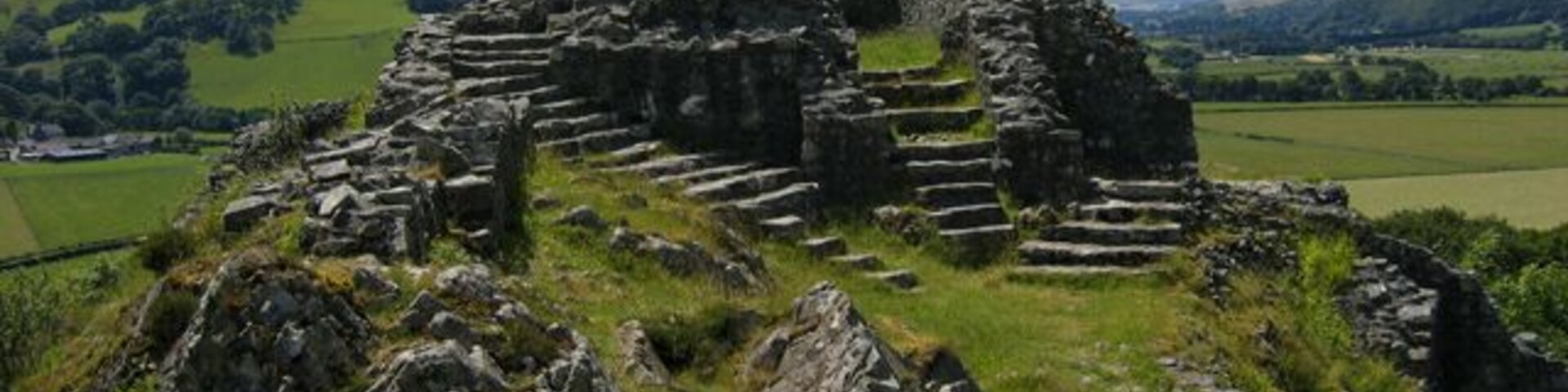 Ruinas de piedra antiguas con estructuras escalonadas y un gran muro de piedra, sobre un fondo de colinas ondulantes y cielo despejado.