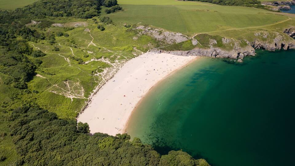 An aerial view of a beach with clear waters, surrounded by green hills and a rocky coastline.