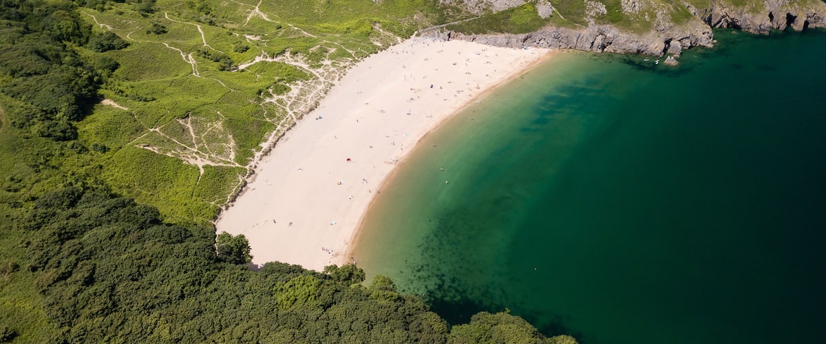 An aerial view of a beach with clear waters, surrounded by green hills and a rocky coastline.