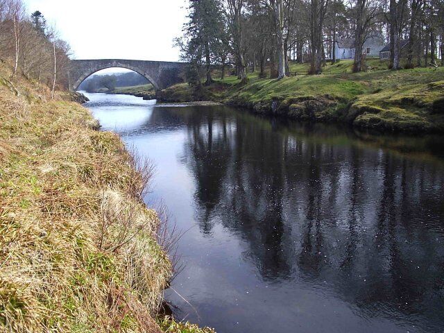 A calm river with a stone bridge, grassy banks, and trees reflecting on the water.