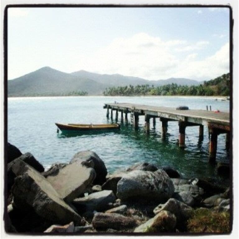 A rocky shoreline with a wooden pier extending into calm water, a small boat docked at the pier, and a mountain range in the background.
