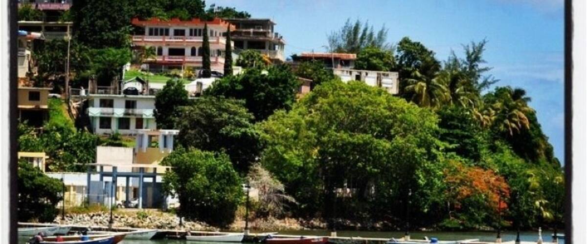 A coastal town with buildings on a hillside, boats on calm water, and lush greenery.