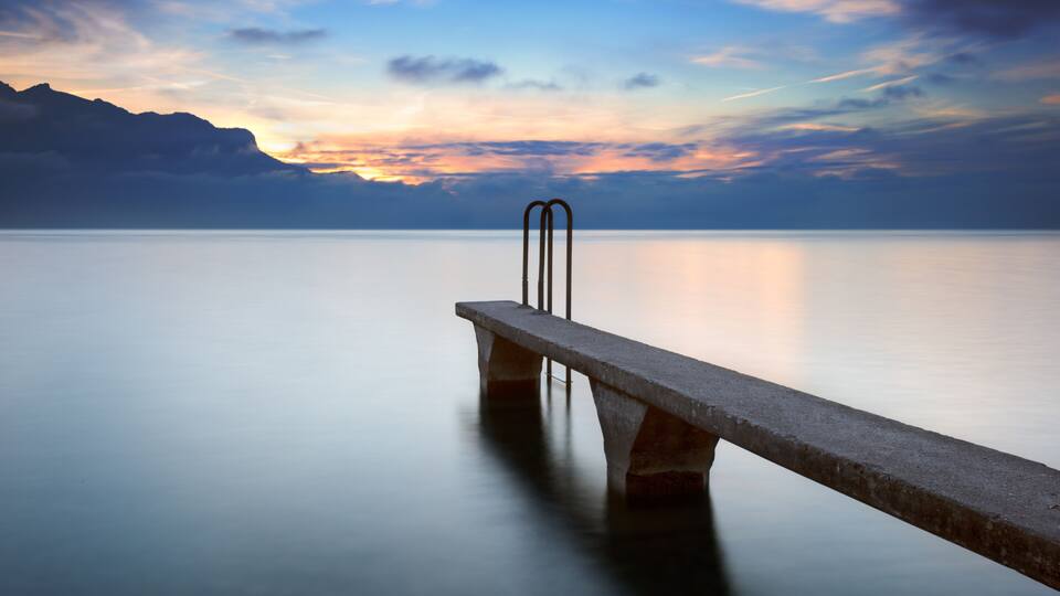 A calm lake with a concrete pier extending into the water, surrounded by mountains and a colorful sunset sky.