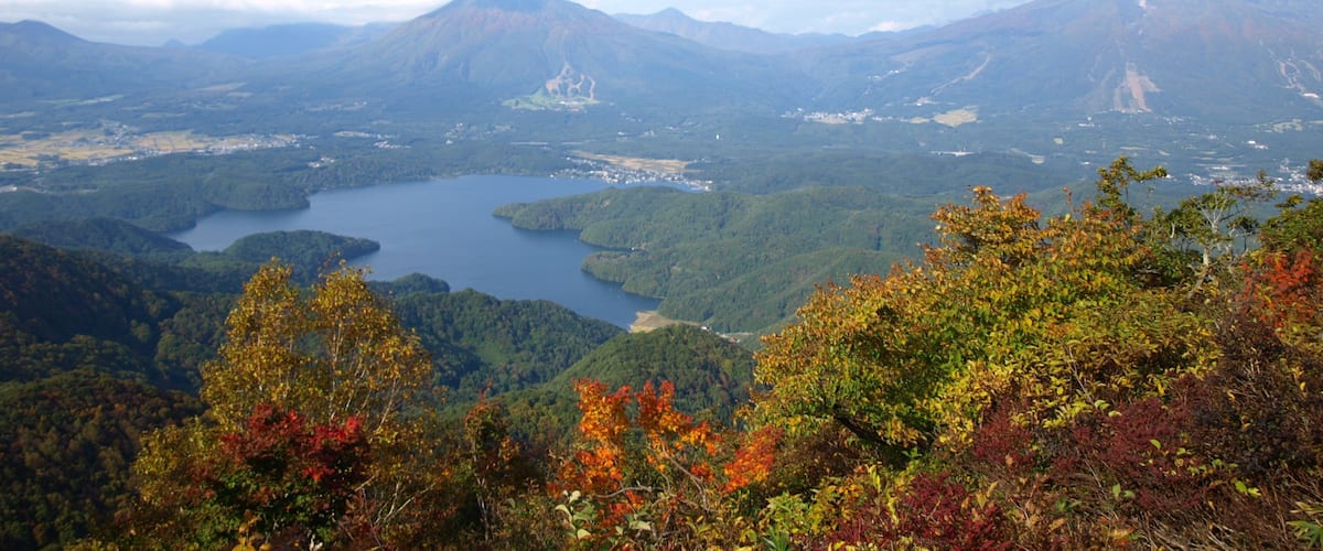 A mountainous landscape with a lake surrounded by dense forests and autumn-colored foliage.