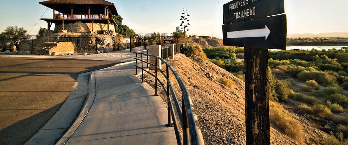 A prisoner's pass trailhead sign, a wooden structure, and a paved path leading to a fenced area.