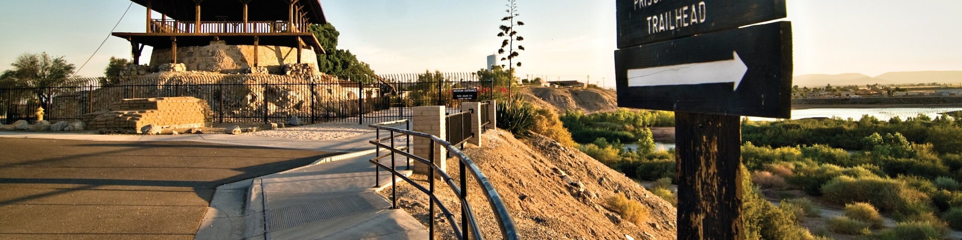 A prisoner's pass trailhead sign, a wooden structure, and a paved path leading to a fenced area.