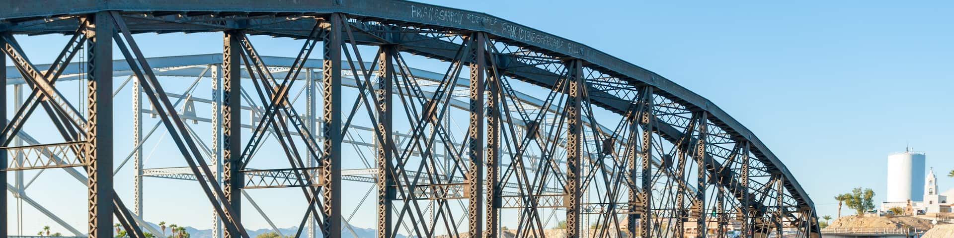 A steel bridge over a river with greenery and a distant cityscape.