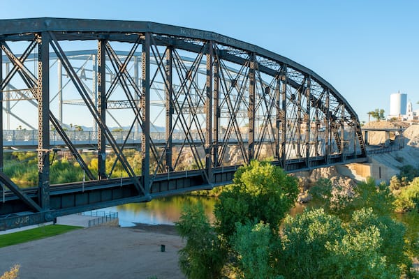 A steel bridge over a river with greenery and a distant cityscape.