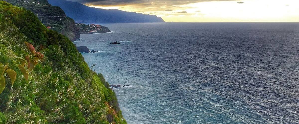 A coastal landscape with a cliff, ocean, and distant mountains under a cloudy sky.