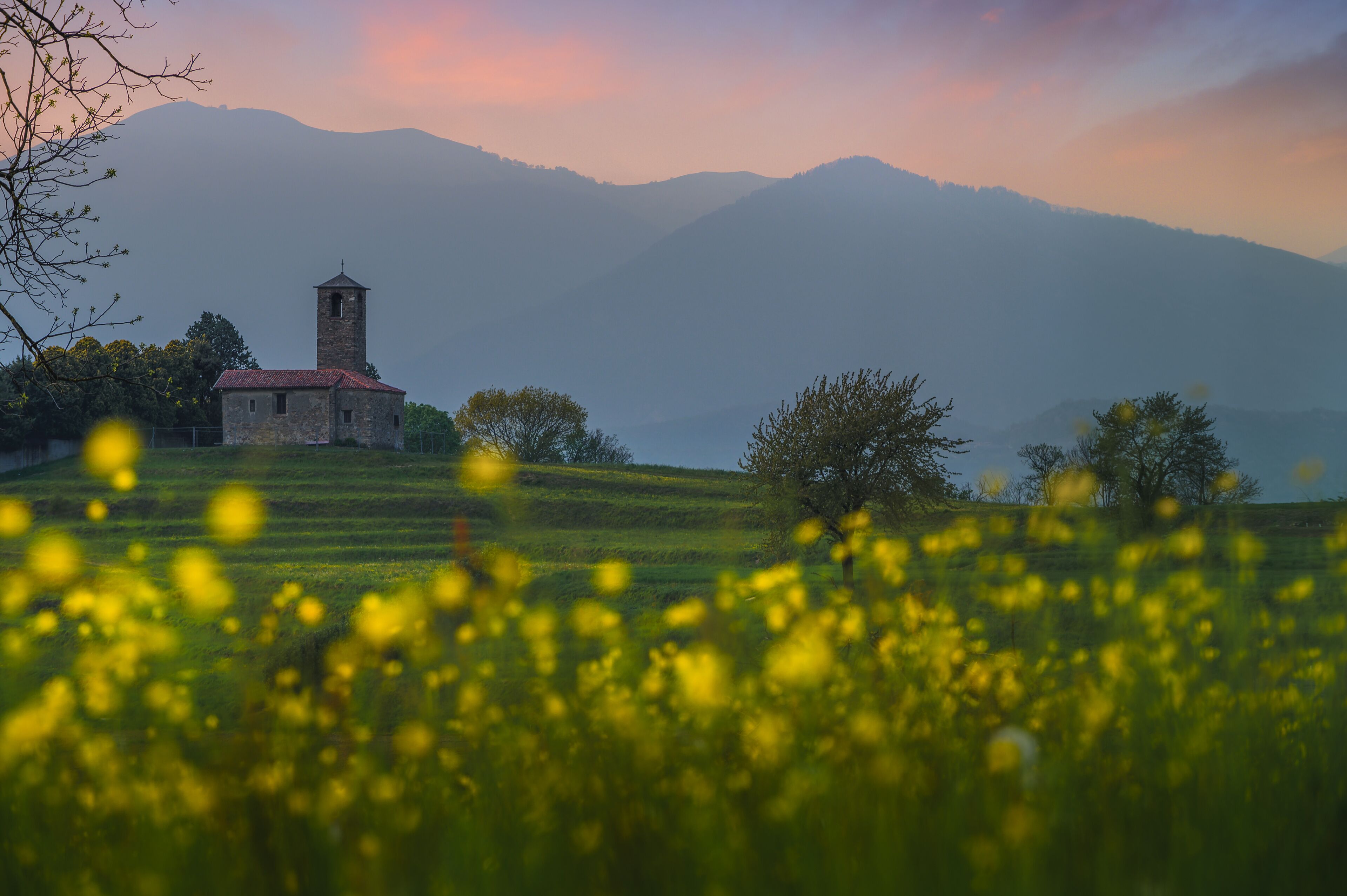 A church with a bell tower on a hill, surrounded by green fields and mountains in the background.