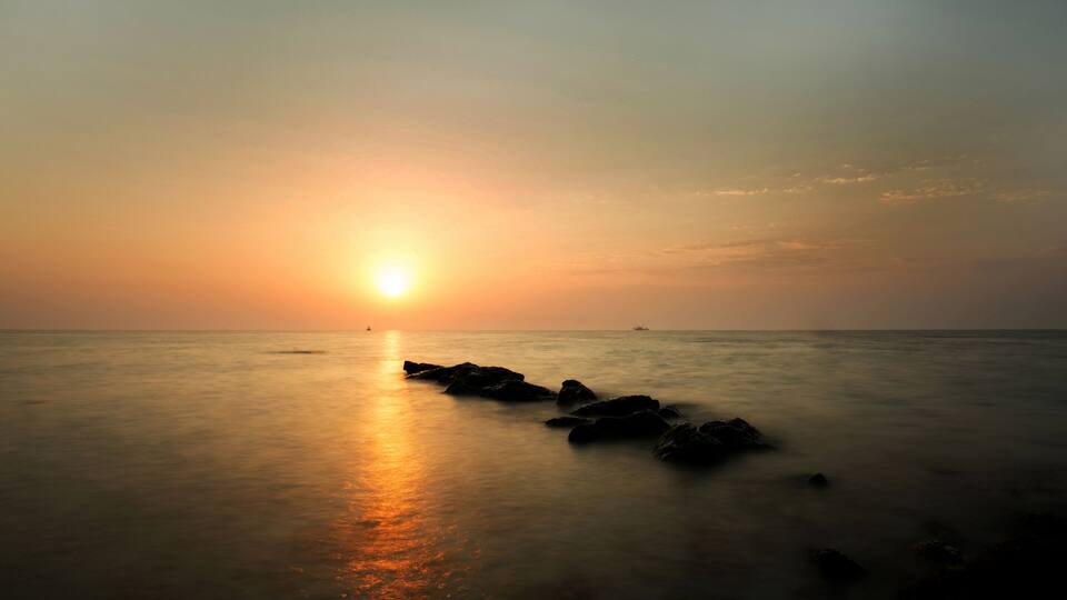 A sunset over calm ocean waters with rocks in the foreground.