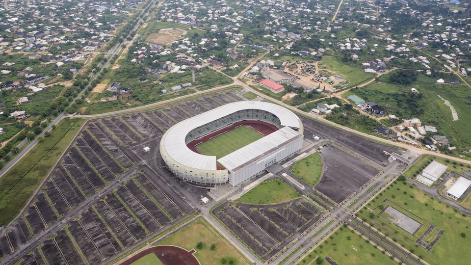 Aerial view of a stadium surrounded by a vast parking area and green spaces blending into the city's edge, Akwa Ibom, Uyo, Nigeria.