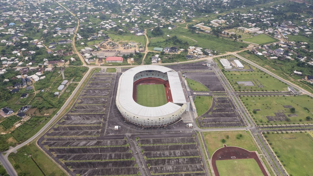Aerial view of the expansive, modern stadium with its oval architecture and surrounding parking lots set against the backdrop of a densely populated urban area, Uyo, Nigeria.