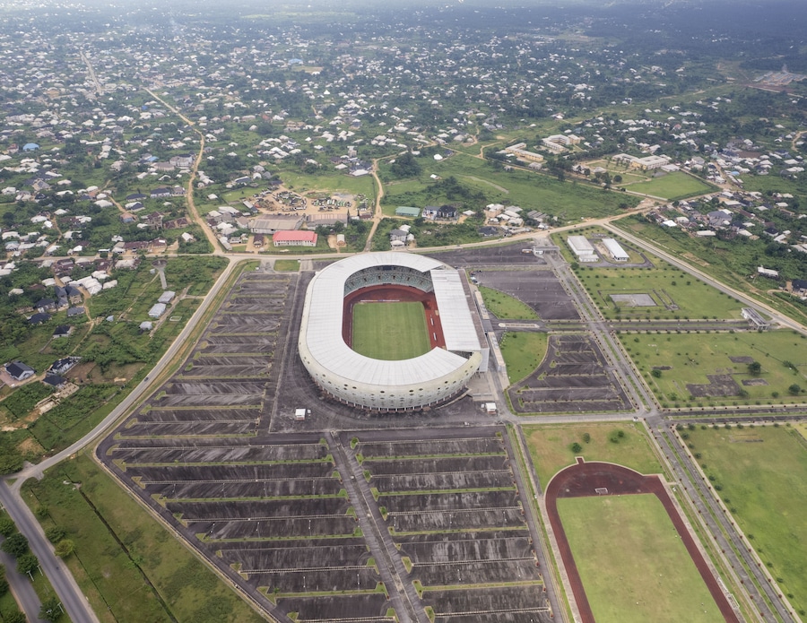 Aerial view of the expansive, modern stadium with its oval architecture and surrounding parking lots set against the backdrop of a densely populated urban area, Uyo, Nigeria.