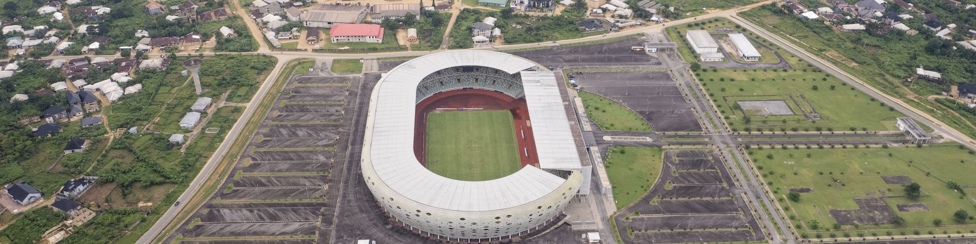 Aerial view of the expansive, modern stadium with its oval architecture and surrounding parking lots set against the backdrop of a densely populated urban area, Uyo, Nigeria.