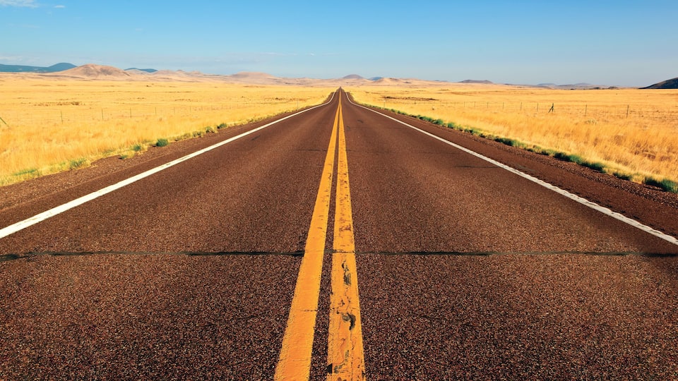 A straight road with yellow centerline, flanked by dry grass fields and distant hills under a clear sky.
