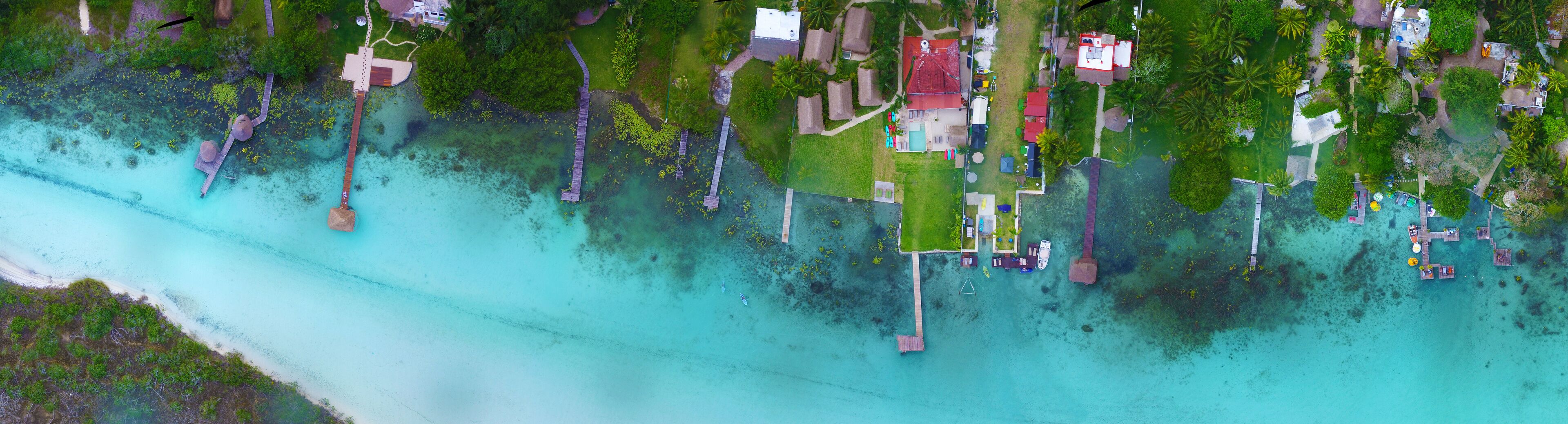An aerial view of a tropical island with clear turquoise waters, a wooden pier, and several thatched-roof huts.