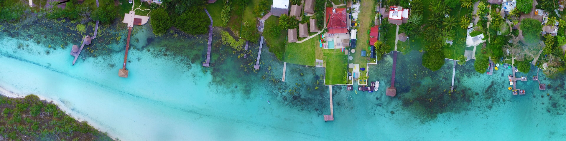 An aerial view of a tropical island with clear turquoise waters, a wooden pier, and several thatched-roof huts.