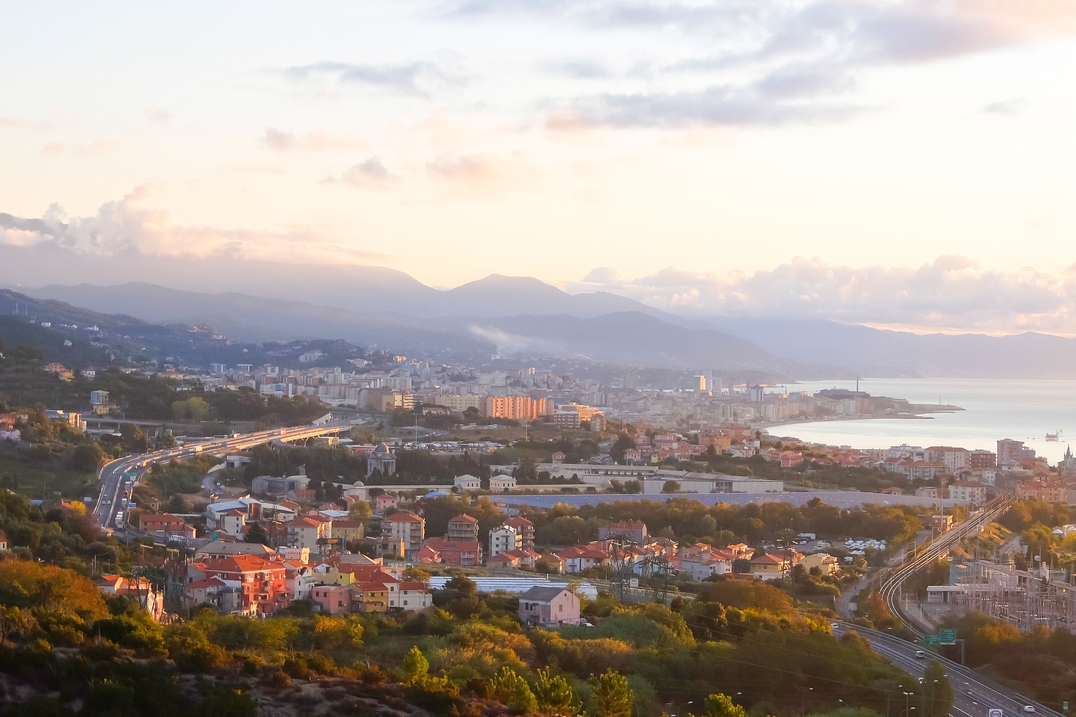 A coastal cityscape with a winding road, residential buildings, and a view of the sea and mountains in the distance.