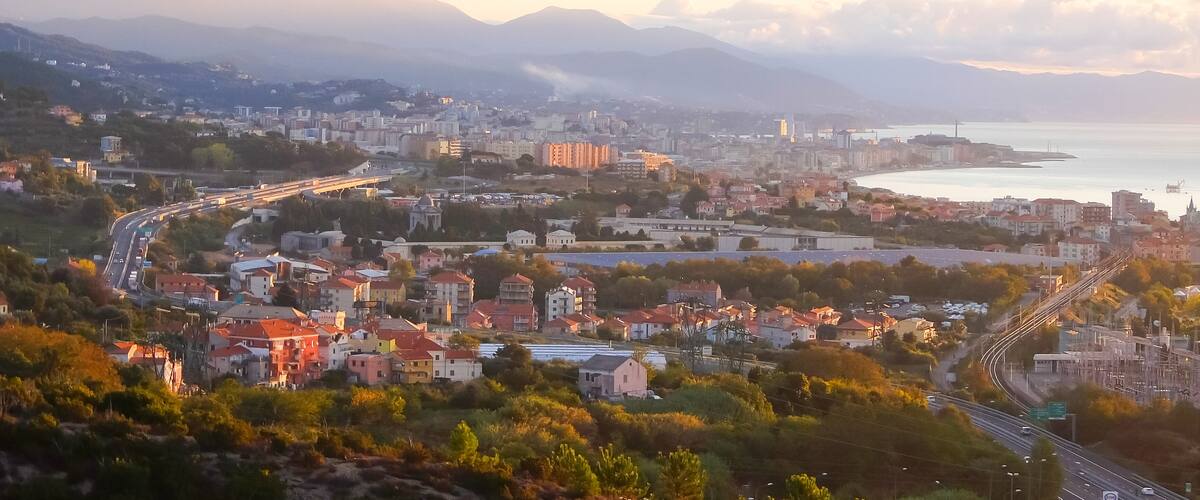 A coastal cityscape with a winding road, residential buildings, and a view of the sea and mountains in the distance.