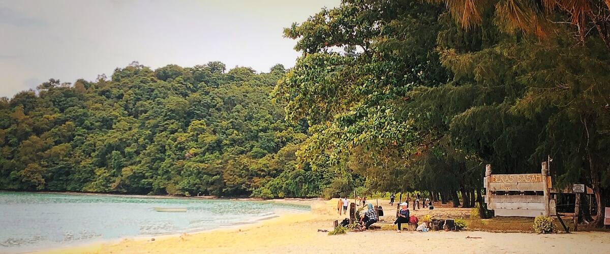 A beach with people relaxing, a sign reading "BERAS BASAH," and a dense forest in the background.