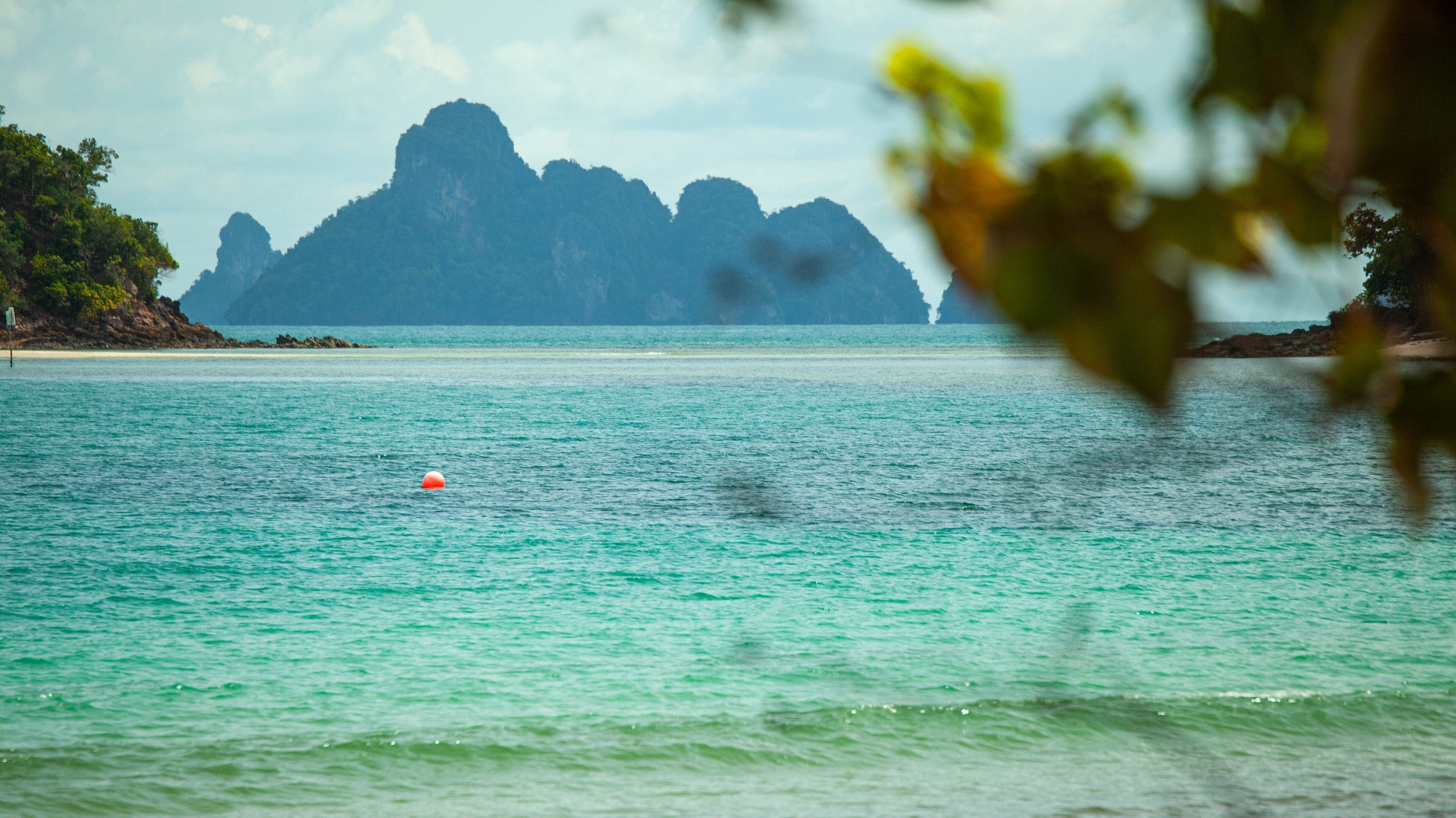 A turquoise sea with a buoy, a beach, and a mountain range in the distance.