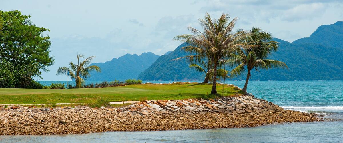 A golf course with a rocky shoreline and palm trees by a lake with mountains in the background.