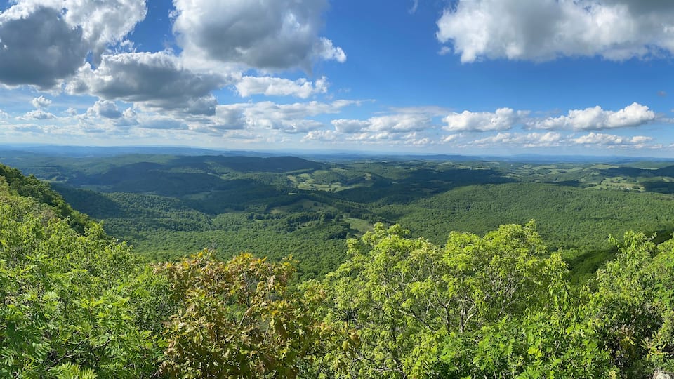 A mountainous landscape with dense green forests and a clear blue sky with scattered clouds.