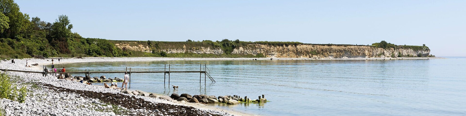 A rocky shoreline with a pier extending into calm waters, surrounded by cliffs and greenery.